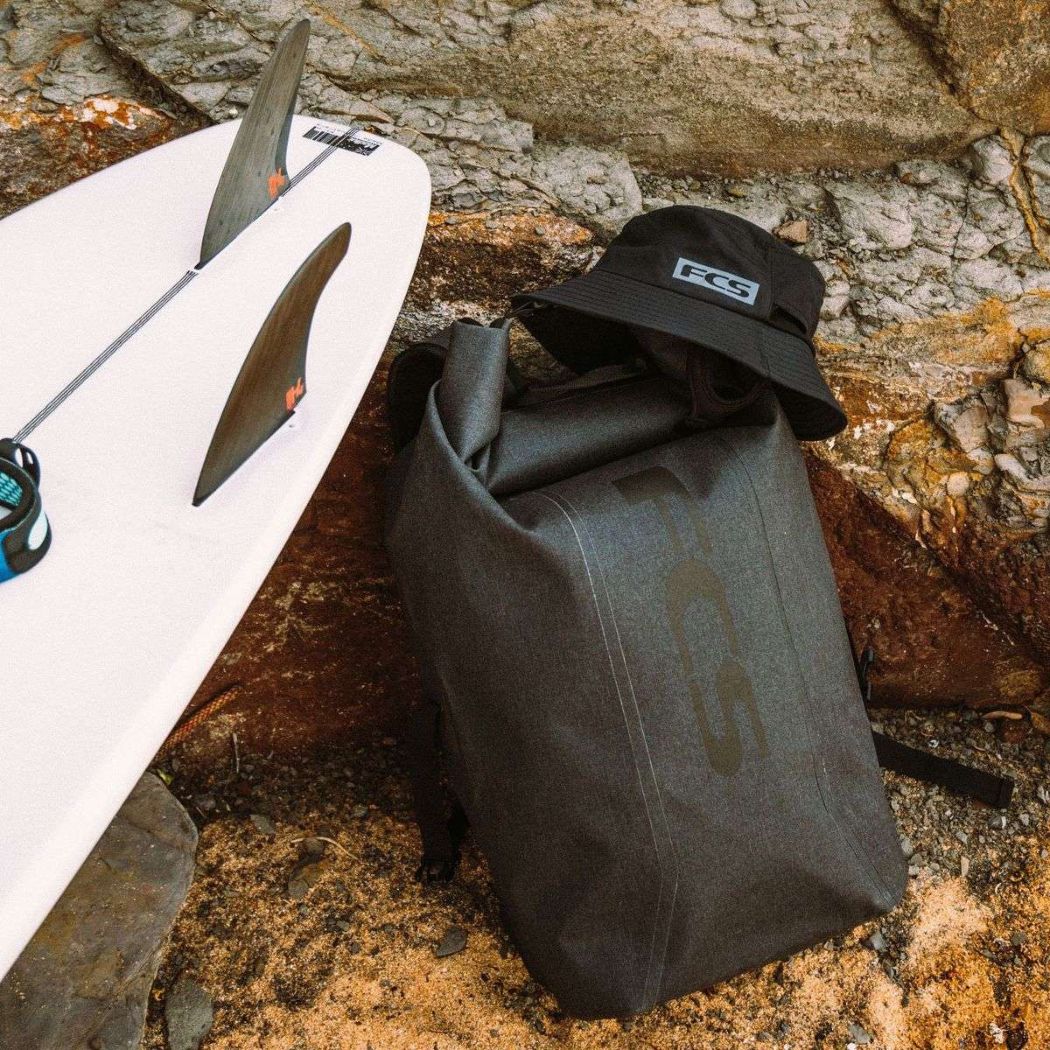 FCS Dry-Wet Rucksack mit einem Surfboard und FCS Buckethat vor Steinen