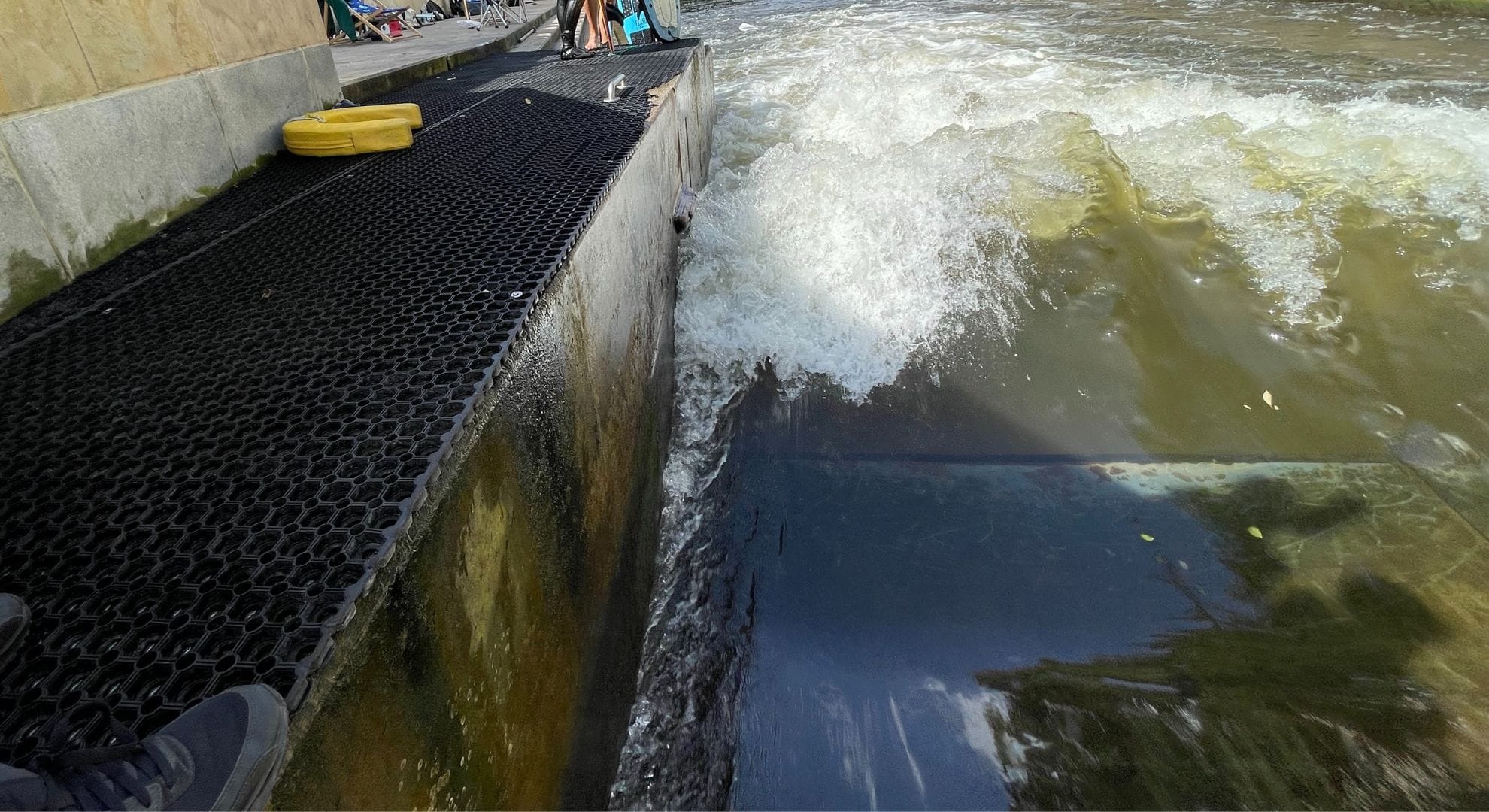 Eine schwarze Metallplattform befindet sich über dem aufgewühlten Flusswasser neben einer Betonwand. Ein gelber Schwimmer und ein Paar Schuhe befinden sich auf der Plattform. Das Wasser fließt schnell und erzeugt Schaum und Wellen.