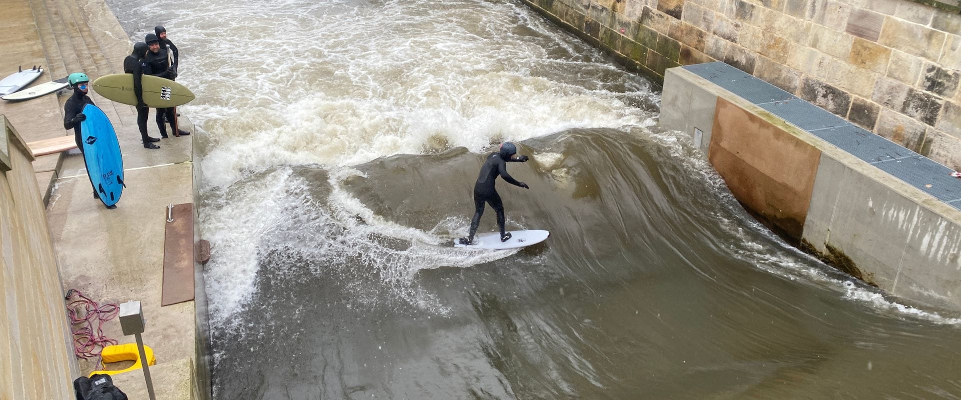 Eine Person surft auf einer künstlichen Flusswelle in der Nähe einer Steinmauer, während drei Personen in Neoprenanzügen mit Surfbrettern an der Seite stehen und zusehen. Das Wasser scheint turbulent zu sein.