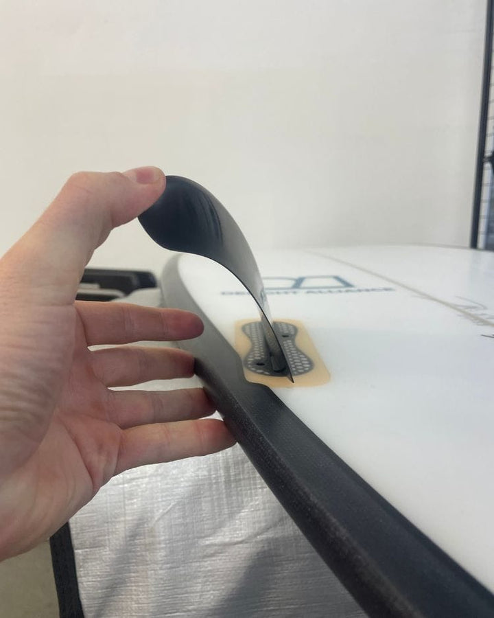 Eine Hand drückt auf eine biegsame schwarze Flosse, die an einem weißen Surfbrett befestigt ist, wodurch sich die Flosse zur Seite biegt. Das Brett liegt in einem Innenraum in der Nähe einer Drahtgitterwand.