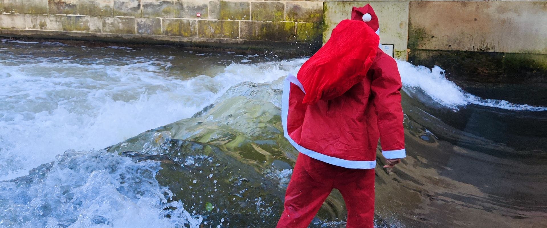 Eine Person in einem roten Weihnachtsmannkostüm steht am Rande eines schnell fließenden, flachen Flusses mit weißem Wasser in der Nähe einer Betonmauer.