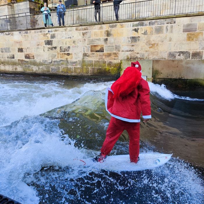 Eine Person in einem roten Weihnachtsmann-Outfit surft auf einer künstlichen Welle in einem Fluss, während mehrere Personen hinter einer Steinmauer darüber zusehen.