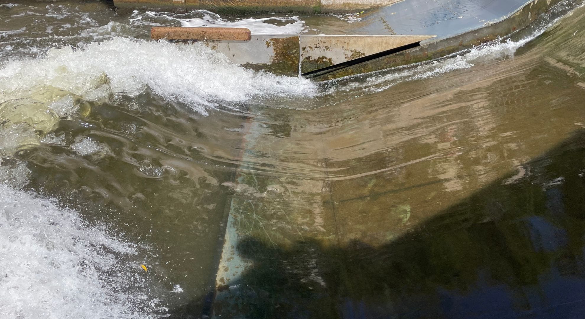 Wasser fließt über eine schräge Metallstruktur und erzeugt weißen Schaum und Wellen. Ein brauner Gegenstand, möglicherweise ein Ast oder ein Stock, ruht auf der Oberkante des Metalls. Auf der Wasseroberfläche sind Spiegelungen und Schatten zu sehen.
