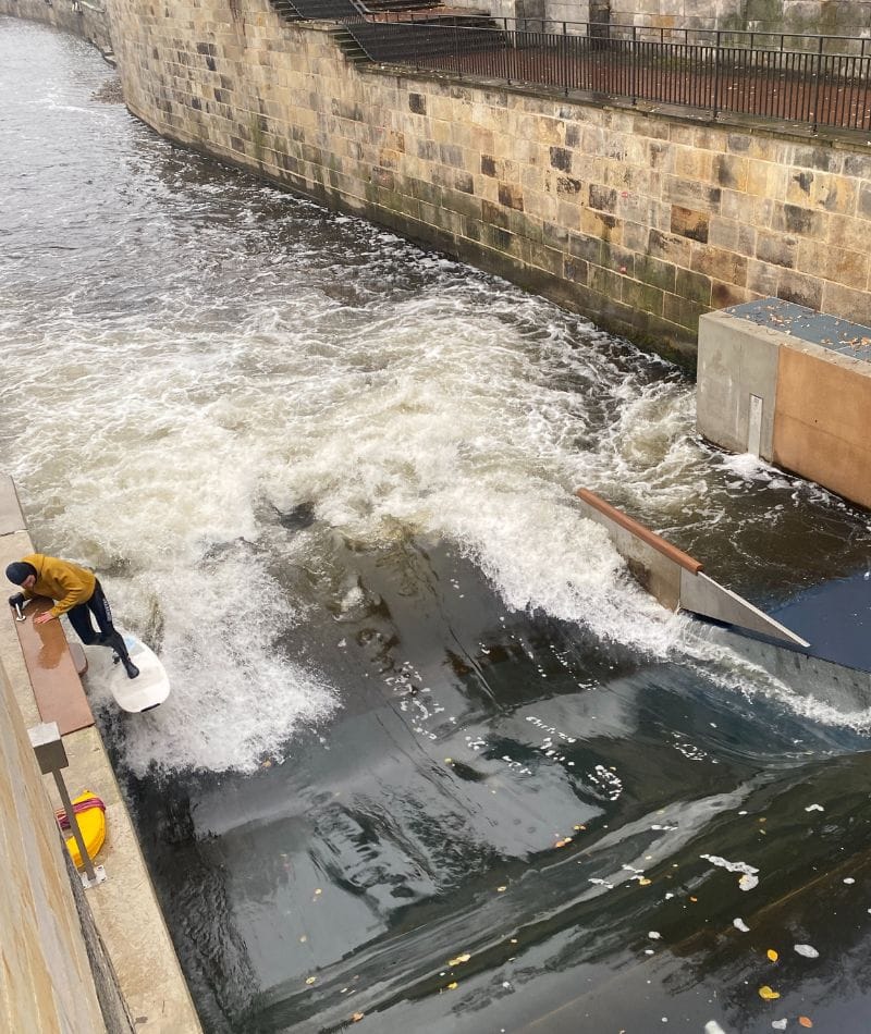 Eine Person bereitet sich darauf vor, auf ein weißes Surfbrett zu steigen, das am Rande eines Betonsockels neben einer künstlichen Flusswelle steht, die von Steinmauern gesäumt wird.