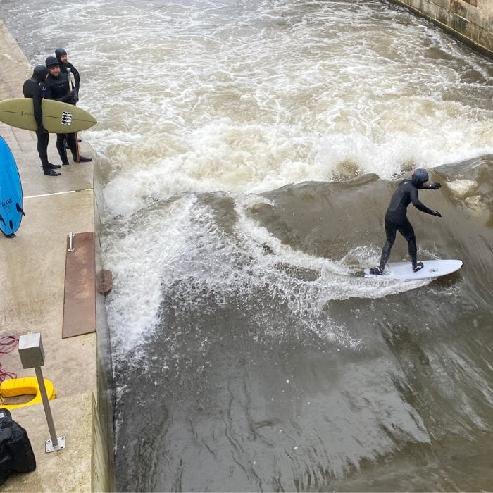 Eine Person in einem Neoprenanzug surft auf einer stehenden Welle in einem Betonkanal, während zwei andere Personen mit Surfbrettern vom Rand aus zusehen. Das Wasser ist unruhig, und das Wetter scheint bedeckt zu sein. Die Ausrüstung liegt in der Nähe auf dem Boden.