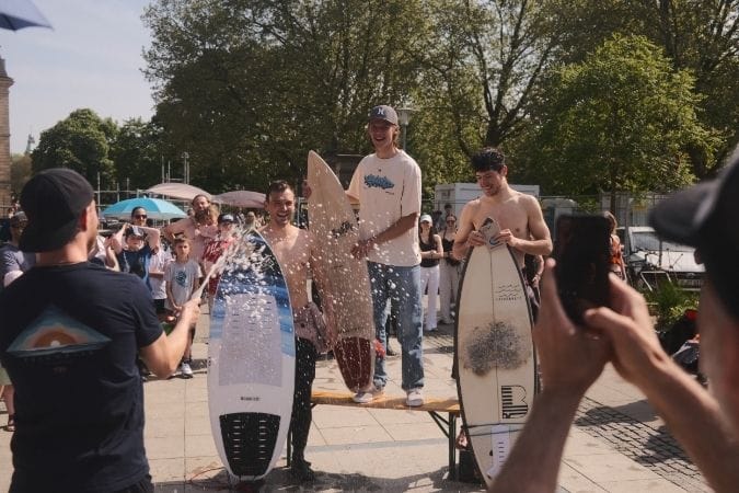 Drei lächelnde Männer mit Surfbrettern stehen auf einer Plattform im Freien, wobei ein Mann Champagner verspritzt, während die Menschen im Hintergrund zusehen und Fotos machen. Hinter der Menge sind Bäume und Sonnenschirme zu sehen.