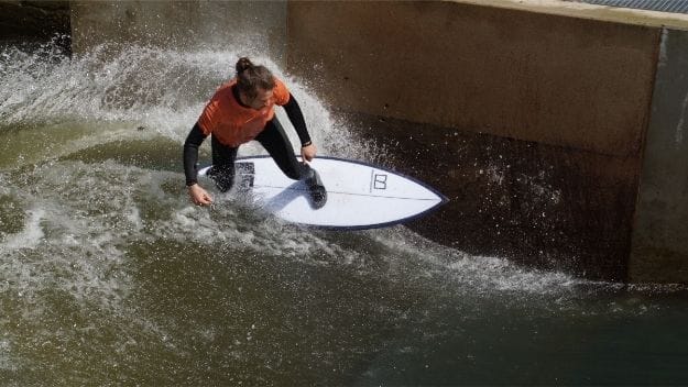 Eine Person in orangefarbenem Hemd und schwarzem Neoprenanzug surft auf einer künstlichen Welle in der Nähe einer Betonmauer, wobei das Wasser um sie herum spritzt.