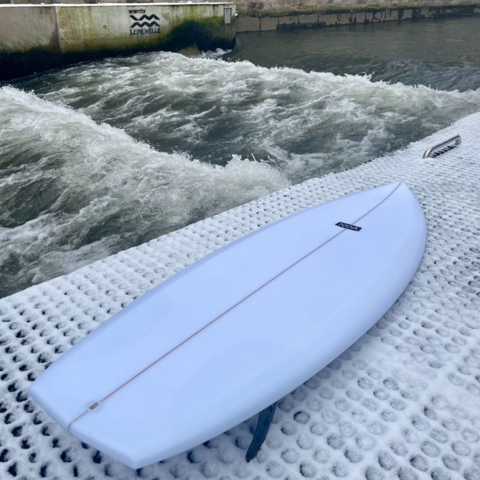 Ein weißes Surfbrett liegt auf einer strukturierten weißen Plattform neben einem künstlichen Wellenbad mit aufgewühltem Wasser. Ein kleines Schild mit der Aufschrift "Leinewelle" ist im Hintergrund an einer Betonwand zu sehen.