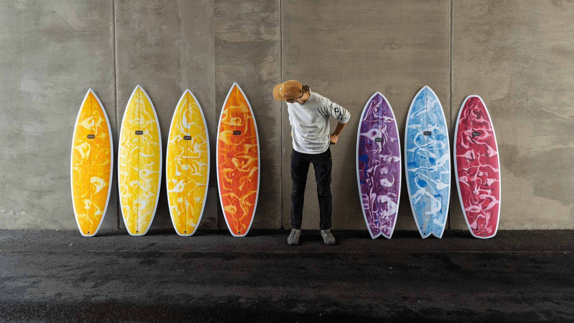 7 Noa Surfboard in verschiedenen Farben an eine Wand gelehnt mit einer Person daneben