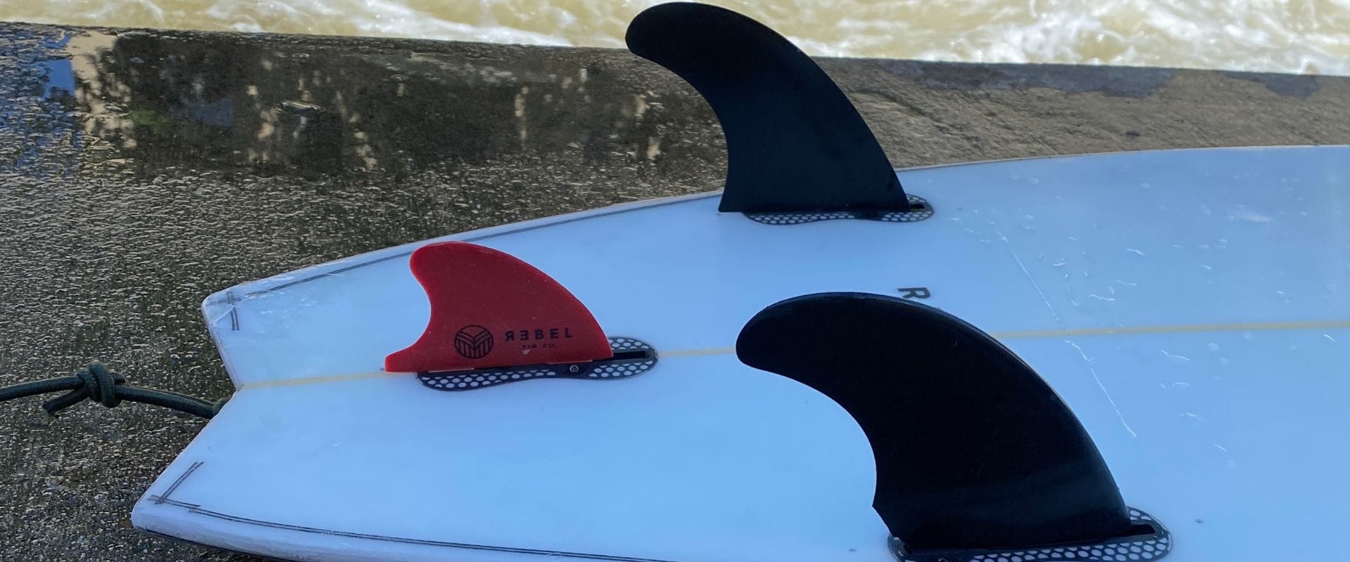 Nahaufnahme eines weißen Surfbretts mit drei Flossen; zwei schwarze Flossen und eine rote Flosse. Auf der roten Flosse sind REBEL und ein Logo aufgedruckt. Das Surfbrett steht auf einer Betonfläche in der Nähe des Wassers.