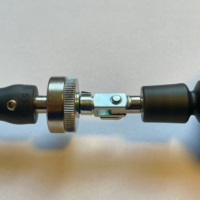 Nahaufnahme eines mechanischen Gelenks aus Metall mit einem gerändelten Einstellring, der zwei schwarze zylindrische Griffe verbindet, die auf einer hellen Fläche liegen.