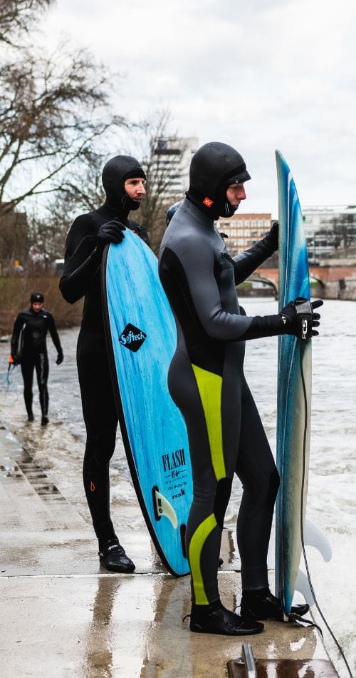Zwei Personen in Neoprenanzügen und Kapuzen stehen an einem Betonufer und halten jeweils ein Surfbrett. Eine dritte Person in einem Neoprenanzug geht auf das Wasser im Hintergrund zu. Auf der anderen Seite des Flusses sind Bäume und Gebäude zu sehen.
