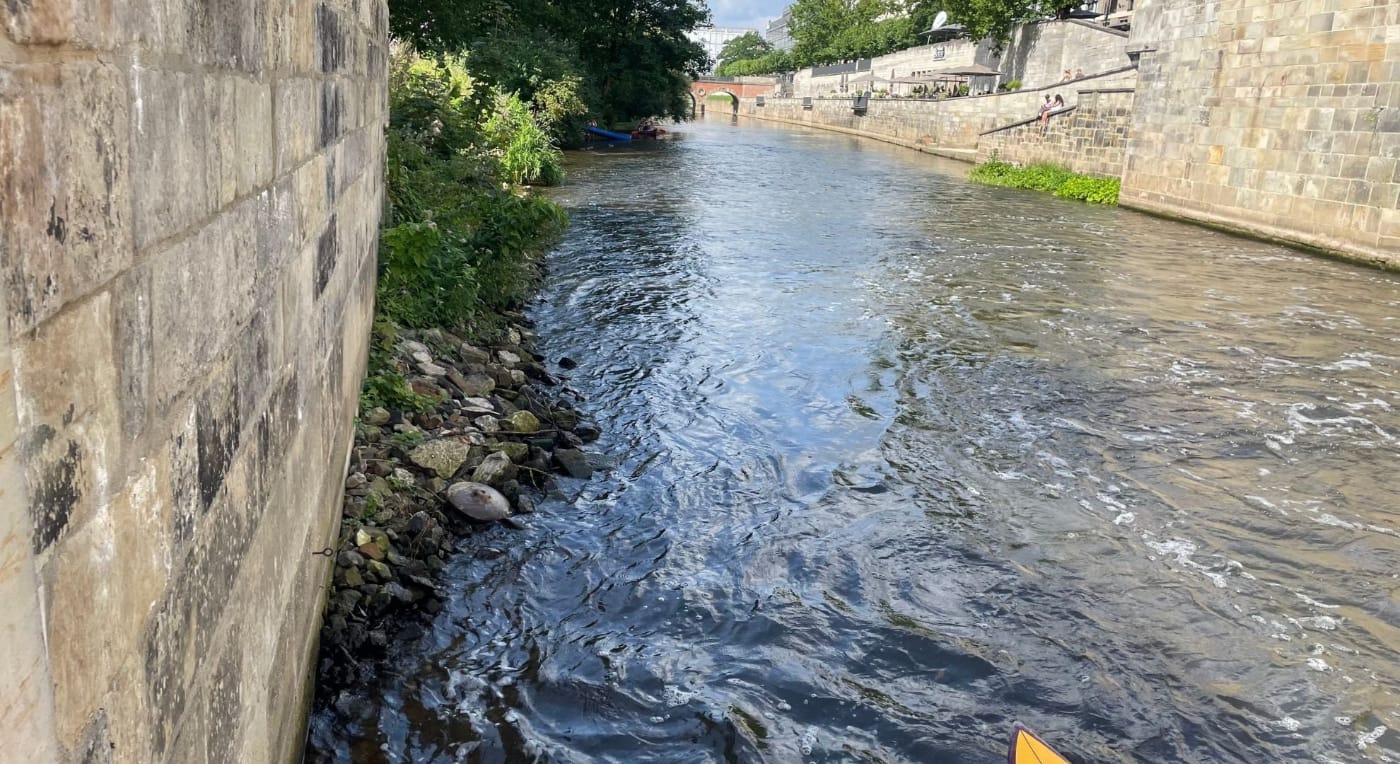 Ein schmaler Fluss fließt zwischen Steinmauern, mit Bäumen und Vegetation an einem Ufer und einer kleinen Brücke in der Ferne unter einem teilweise bewölkten Himmel.