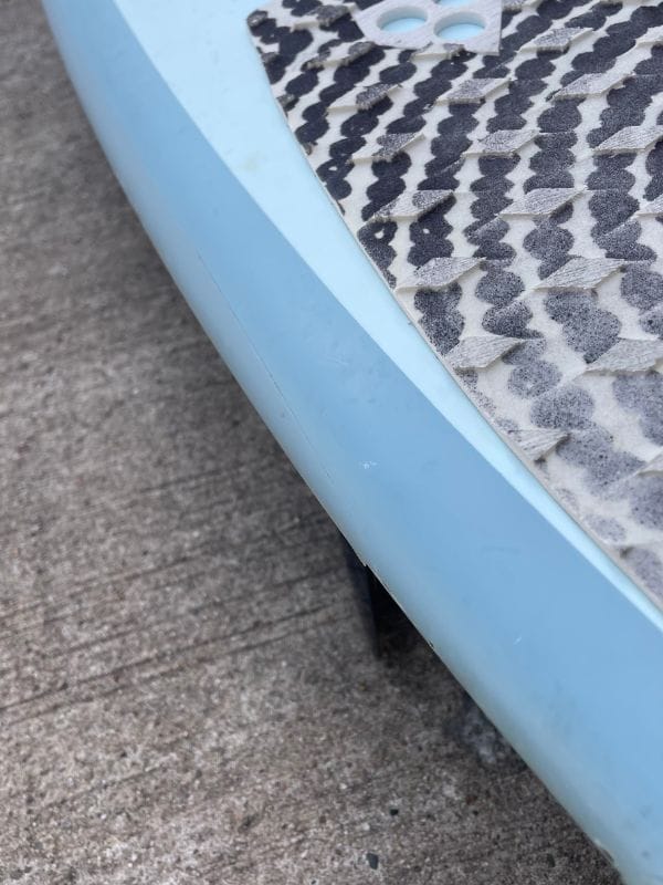 Nahaufnahme der Kante eines hellblauen Paddelboards mit einem strukturierten, schwarz-weißen Deckpad, das auf einer rauen Betonoberfläche ruht.