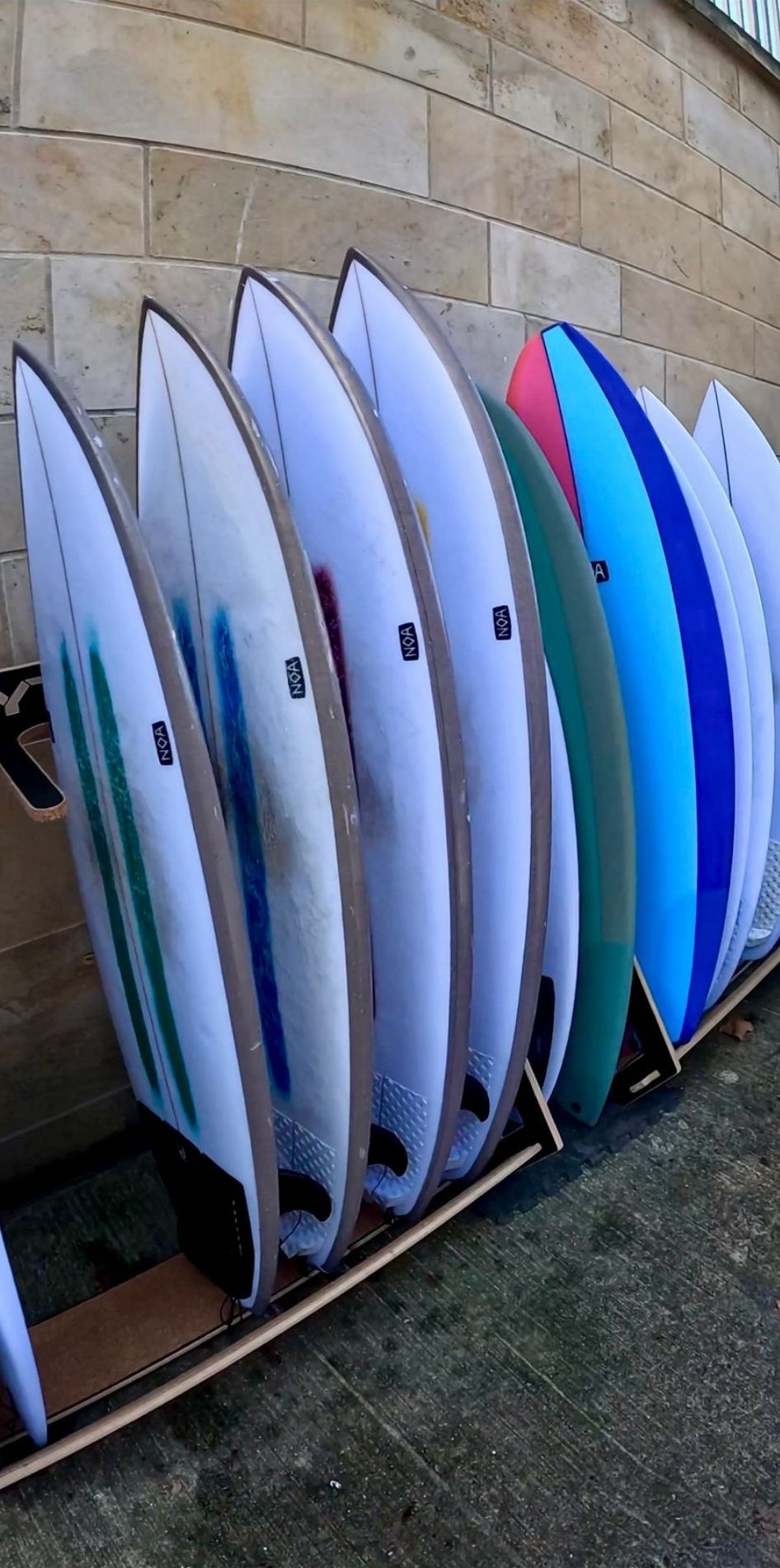Surfboards der Marke NOA stehen aufrecht in einem Surfboardständer vor einer Wand