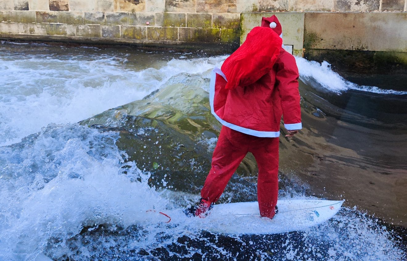 Eine Person, die ein rotes Weihnachtsmannkostüm trägt, surft auf einer Flusswelle in der Nähe einer Steinmauer. Das Foto ist von hinten aufgenommen und zeigt Wasser, das um das Surfbrett herum spritzt.