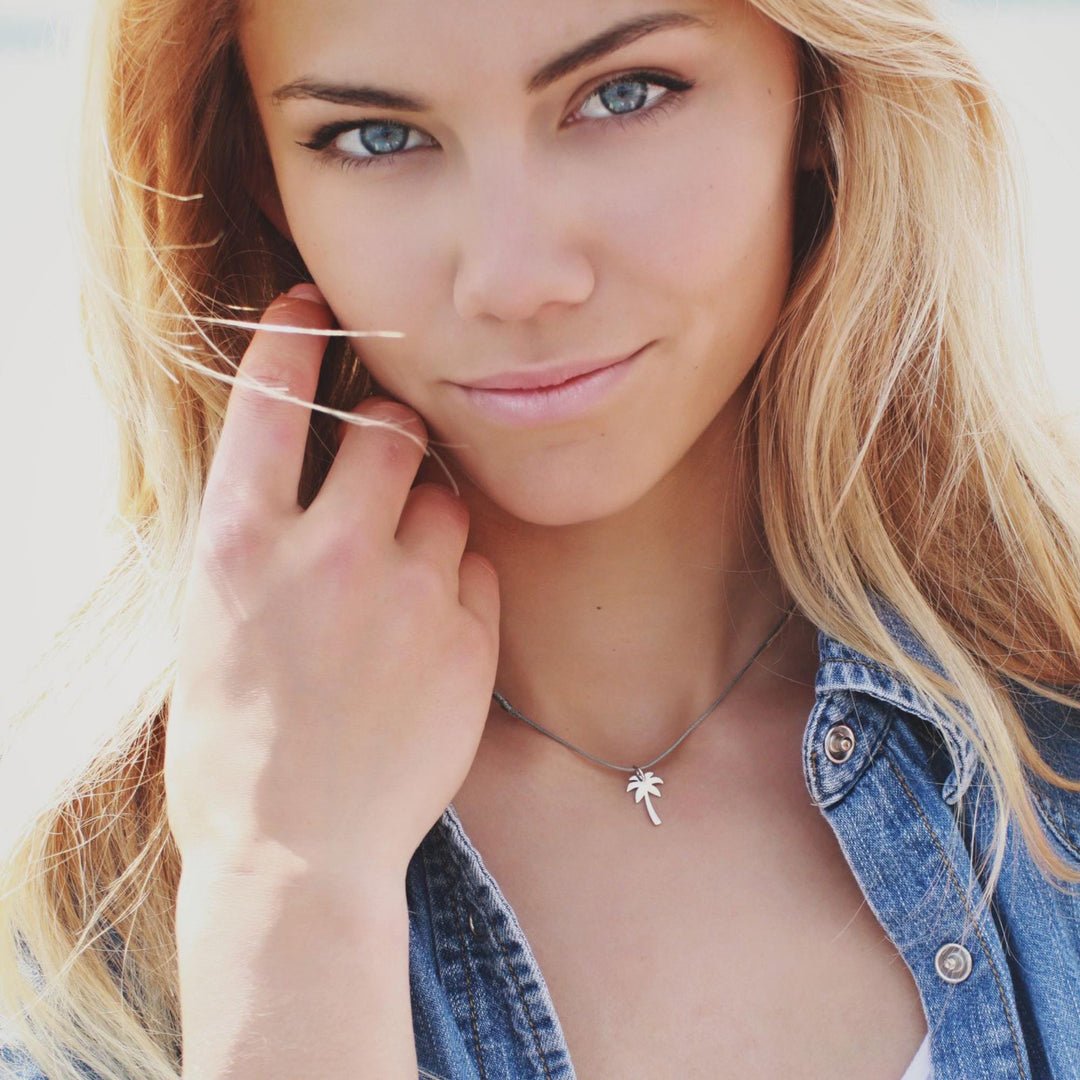 Eine junge Frau mit langen blonden Haaren, die ein Jeanshemd und den Silver+Surf Silber Schmuck Palme Anhänger trägt, schaut in die Kamera und berührt ihr Gesicht. Der helle Hintergrund ist unscharf.