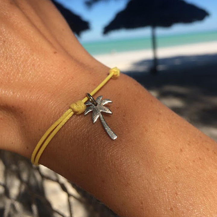 Eine Nahaufnahme eines Handgelenks mit dem Armband Silver+Surf Silber Schmuck Palme von Silver+Surf, mit einer gelben Schnur und einem silbernen Palmenanhänger vor einem Sandstrand, blauem Meer und verschwommenen Sonnenschirmen.