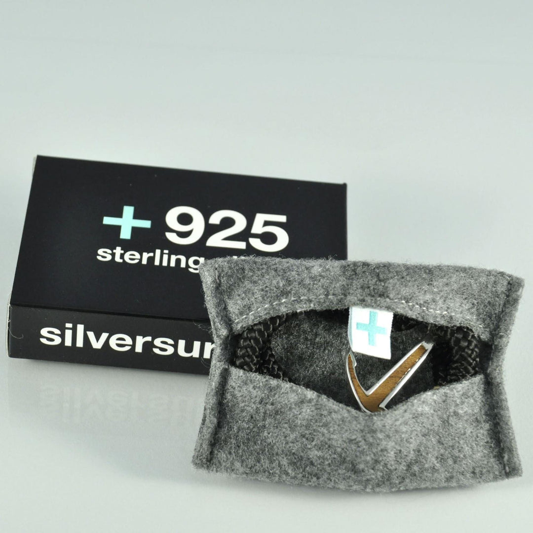 In einem grauen Filzbeutel befindet sich ein silberner Ring mit V-förmigem Muster. Dahinter vervollständigt eine schwarze Schachtel mit der Aufschrift Silver+Surf Silber Schmuck Boot Anker vergoldet die elegante Präsentation.