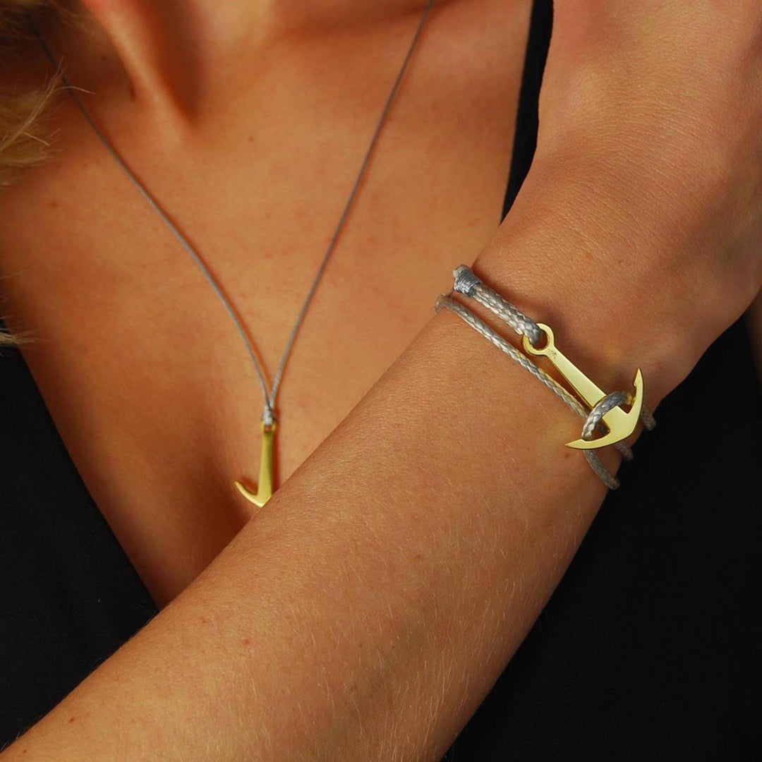 Eine Person in einem schwarzen Oberteil trägt den Silver+Surf Silber Schmuck Boot Anker vergoldet Anhänger Halskette und ein passendes vergoldetes Armband mit grauer Kordel, beide inspiriert von 925 Sterling Silber und entworfen von Silver+Surf.