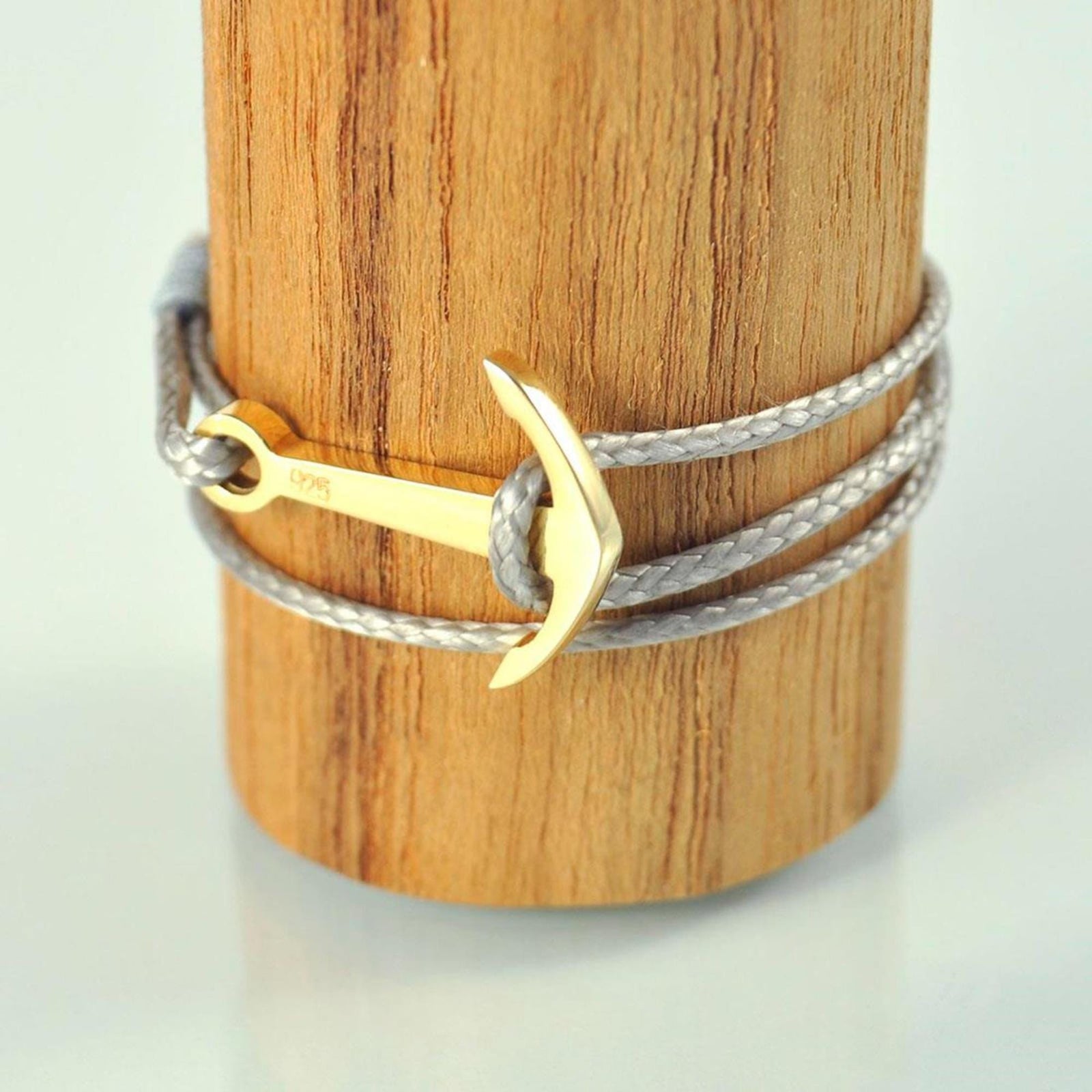 Der Silver+Surf Silber Schmuck Boot Anker vergoldet verfügt über eine goldene ankerförmige Schließe an einer geflochtenen grauen Kordel, die elegant um einen zylindrischen Holzständer gewickelt ist.