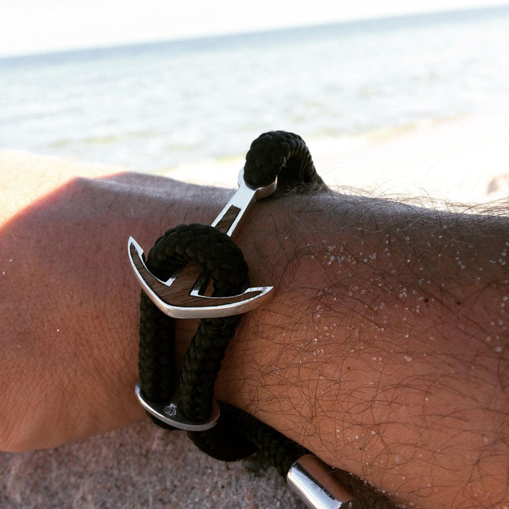 Eine Nahaufnahme eines Handgelenks, das das Silver+Surf Silber Schmuck Boot Anker Wood Armband mit einem schwarzen Seil und einem silbernen Ankeranhänger trägt. Sandkörner liegen auf der Haut, im Hintergrund sind die Küste und das Meer zu sehen.