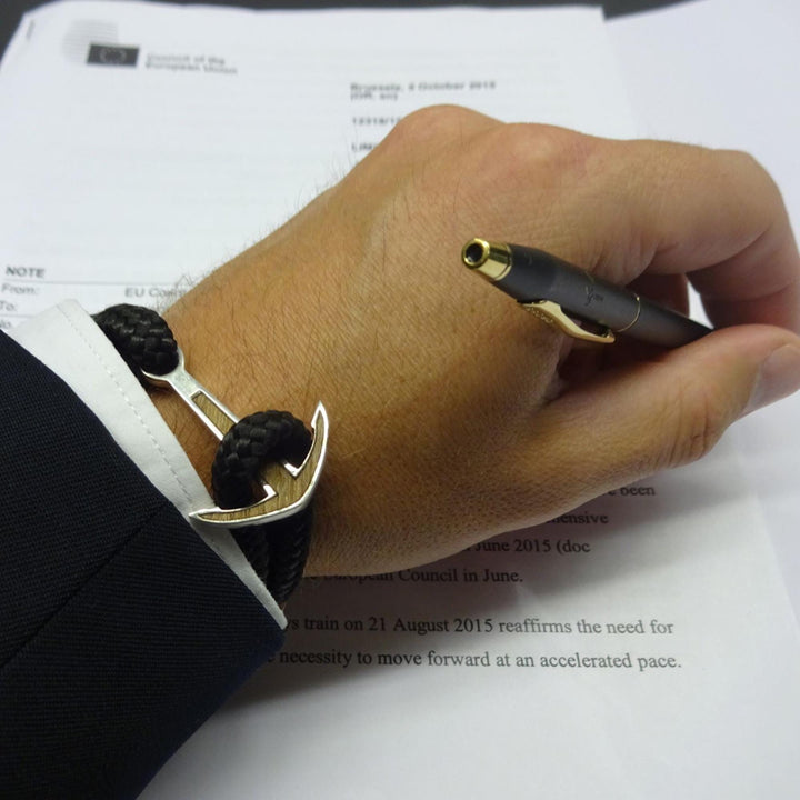 Eine Person im Anzug hält einen Stift über ein Dokument und trägt ein Silver+Surf Silber Schmuck Boot Anker Wood Armband von Silver+Surf an ihrem Handgelenk. Das Dokument hat gedruckten Text und eine offiziell aussehende Kopfzeile.