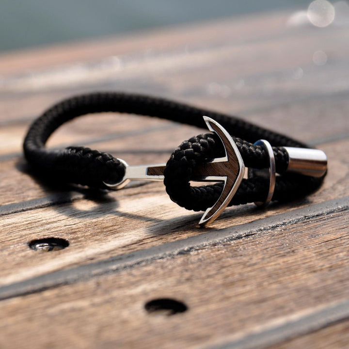 Ein Silver+Surf Silber Schmuck Boot Anker Holz Armband mit einer schwarzen geflochtenen Kordel und Sterling Silber Anker Anhänger ruht auf einer Holzoberfläche mit sichtbaren Schrauben.