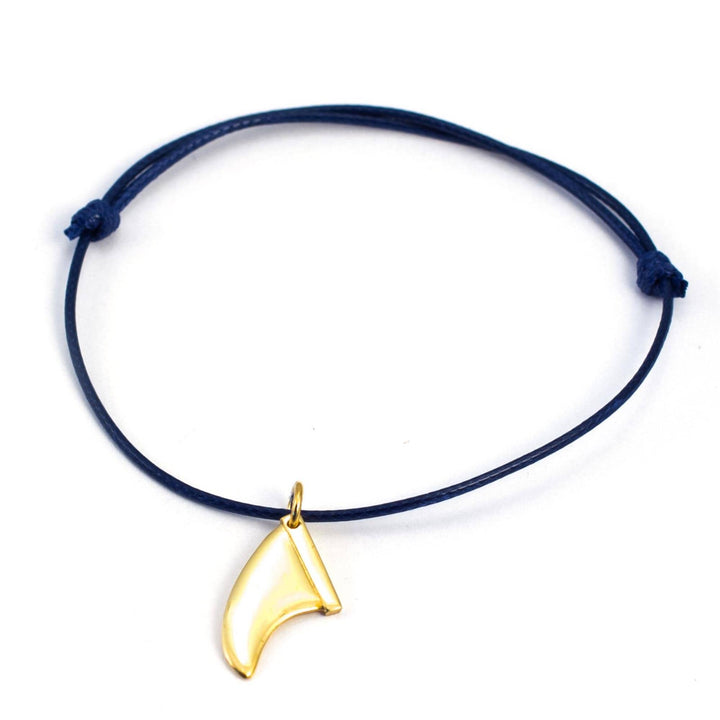 Ein marineblaues Kordelarmband mit einem goldfarbenen Surfboard-Flossen-Charm von Silver+Surf, inspiriert von dem Golddesign der Silber Schmuck Surfboard Finne, auf weißem Hintergrund.