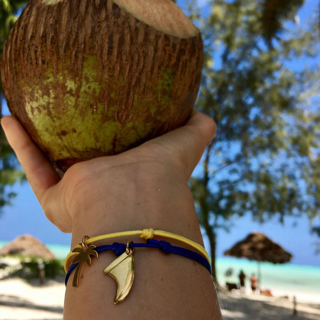 Eine Hand, die Silver+Surf Silber Schmuck Surfboard Finne Goldarmband trägt, hält eine Kokosnuss. Der Hintergrund zeigt einen Sandstrand, Bäume, Sonnenschirme, türkisfarbenes Wasser und einen blauen Himmel.