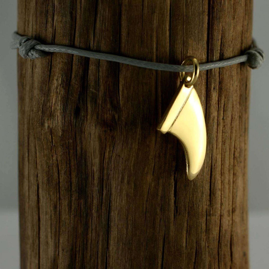 Ein goldener Surfbrett-Flossen-Anhänger von Silver+Surf hängt an einer grauen Schnur, die um verwittertes Holz gewickelt ist.