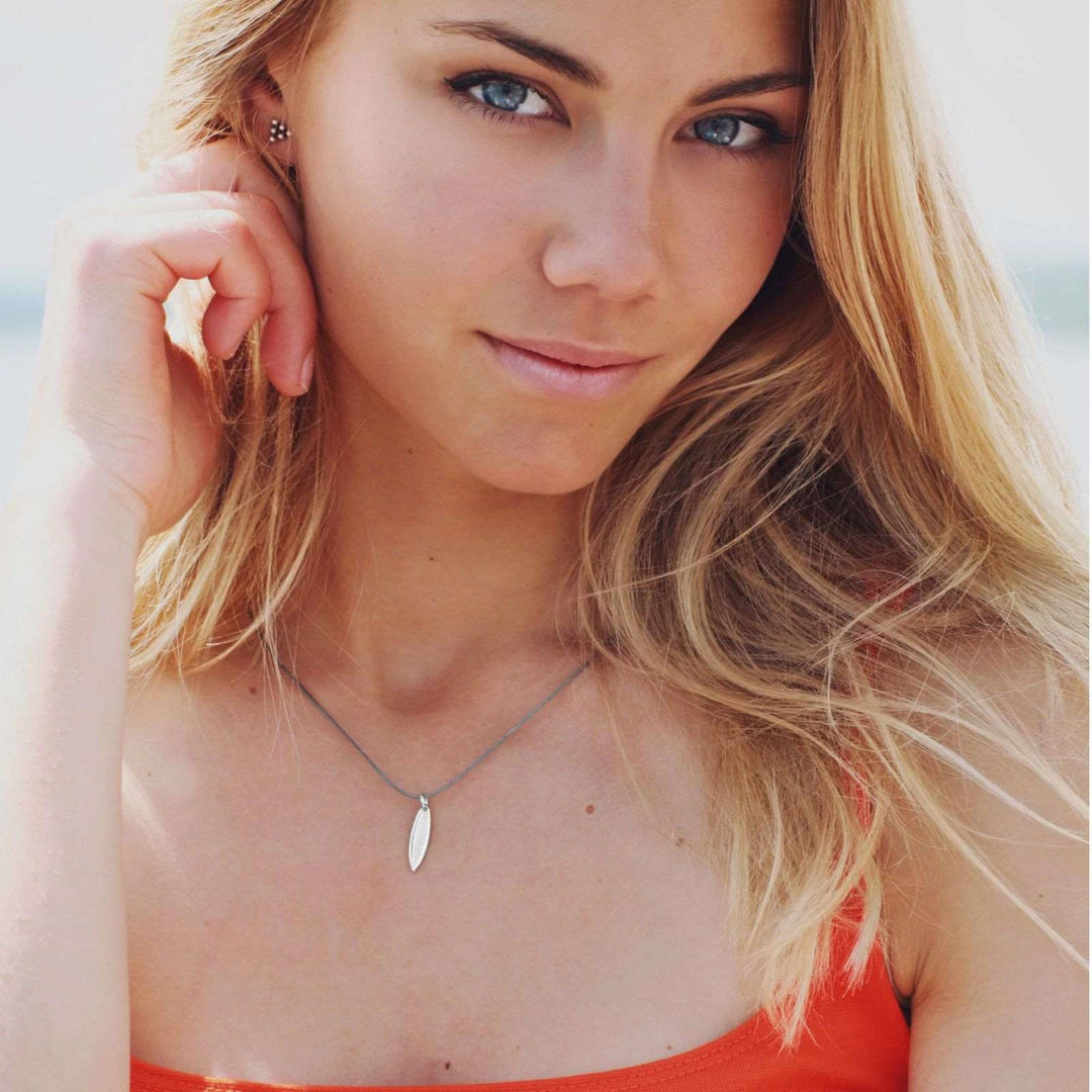 Eine junge Frau mit langen blonden Haaren und blauen Augen trägt ein orangefarbenes Oberteil und die Silver+Surf Silber Schmuck Surfboard Goldkette von Silver+Surf. Sie schaut in die Kamera und hält ihr Haar zurück; der Hintergrund ist unscharf.