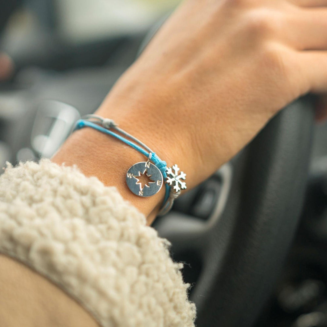 Die Hand einer Person, die ein Lenkrad hält, trägt einen beigen Ärmel und zwei Armbänder: das Silver+Surf Silber Schmuck Windrose Himmelsrichtung von Silver+Surf und ein weiteres mit einer kleinen silbernen Schneeflocke an einer blauen Kordel.
