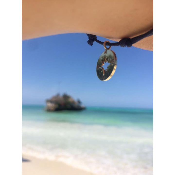 Eine Nahaufnahme zeigt ein Handgelenk, das das Armband Silver+Surf Silber Schmuck Windrose Himmelsrichtung trägt; im unscharfen Hintergrund glitzern eine kleine Felseninsel und das türkisfarbene Meer vor dem blauen Himmel.