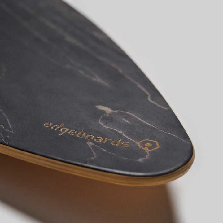 Nahaufnahme der Spitze des dunklen Edgeboards Balanceboards mit Holzkante. Der Schriftzug "Edgeboards" und ein sechseckiges Logo sind in Gold auf die Oberfläche nahe der Spitze gedruckt. Der schlichte helle Hintergrund erinnert an echtes Skatefeeling.