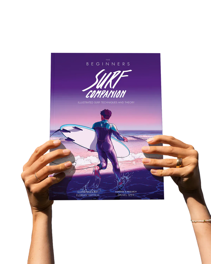 Eine Person hält das Surfcompanion Surf Companion Beginner 2. Edition - Surfbuch, ein illustriertes Arbeitsbuch für Surf-Anfänger, das einen Surfer mit Brett bei Sonnenuntergang zeigt. Beide Hände sind vor einem schlichten weißen Hintergrund zu sehen.