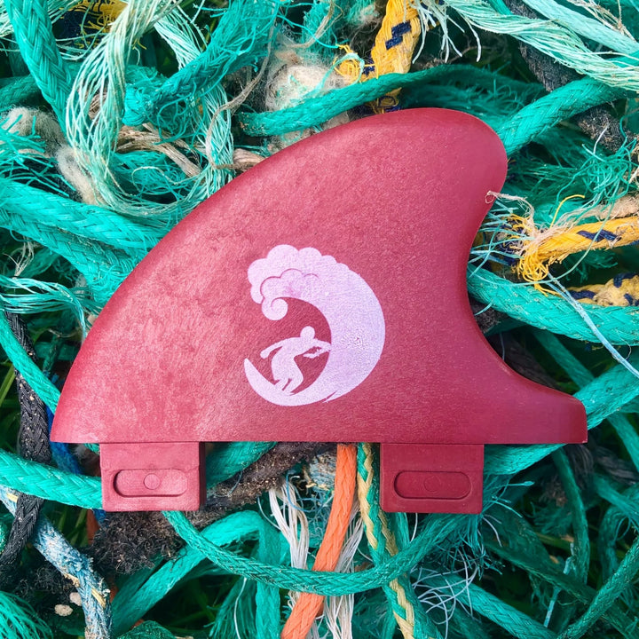 Die Rebel Fin Rebel X Driftwood - Fin Rapid Fin FCS I aus recycelten Fischernetzen hat ein rotes Finish mit einer weißen Wellen- und Surfergrafik, die auf grünen, blauen und orangen Seilen ruht.