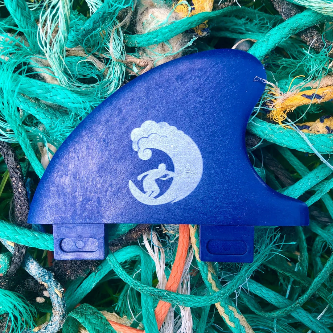 Die Rebel Fin "Rebel X Driftwood - Rapid Fin FCS I" aus recycelten Fischernetzen zeigt eine weiße Surfergrafik auf blauem Grund, die auf grünen, blauen und orangen Seilen ruht - ideal für alle, die nachhaltiges Finnen suchen.