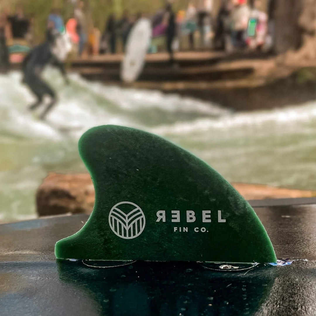 Eine Rebel X Driftwood - Fin Rapid Fin FCS I von Rebel Fin aus recycelten Fischernetzen steht auf nassem Untergrund; im unscharfen Hintergrund surfen Menschen auf einer Flusswelle, umgeben von Bäumen und weiteren Personen.
