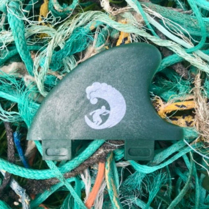 Eine grüne Rebel X Driftwood - Fin Rapid Fin FCS I von Rebel Fin aus recycelten Fischernetzen mit weißem Wellen- und Surfer-Logo liegt auf einer Haufen blauer, grüner und gelber Seile.