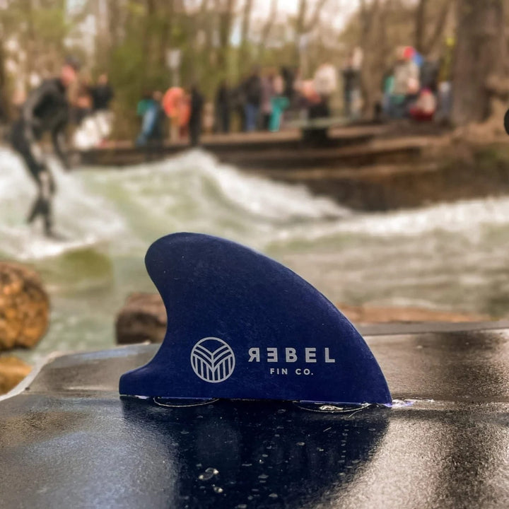 Eine Nahaufnahme einer blauen Rebel Fin Rebel X Driftwood - Fin Rapid Fin FCS I aus recycelten Fischernetzen mit Wassertropfen sitzt auf einer Oberfläche. Im unscharfen Hintergrund sehen Menschen zu, wie jemand auf einem von Bäumen gesäumten Fluss surft.