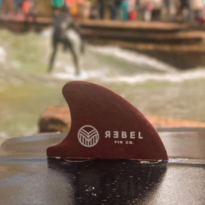 Eine Nahaufnahme einer kastanienbraunen Rebel X Driftwood - Fin Rapid Fin FCS I von Rebel Fin, hergestellt aus recycelten Fischernetzen, auf einer nassen Oberfläche mit einer unscharfen Szene im Freien und Menschen am Wasser im Hintergrund.