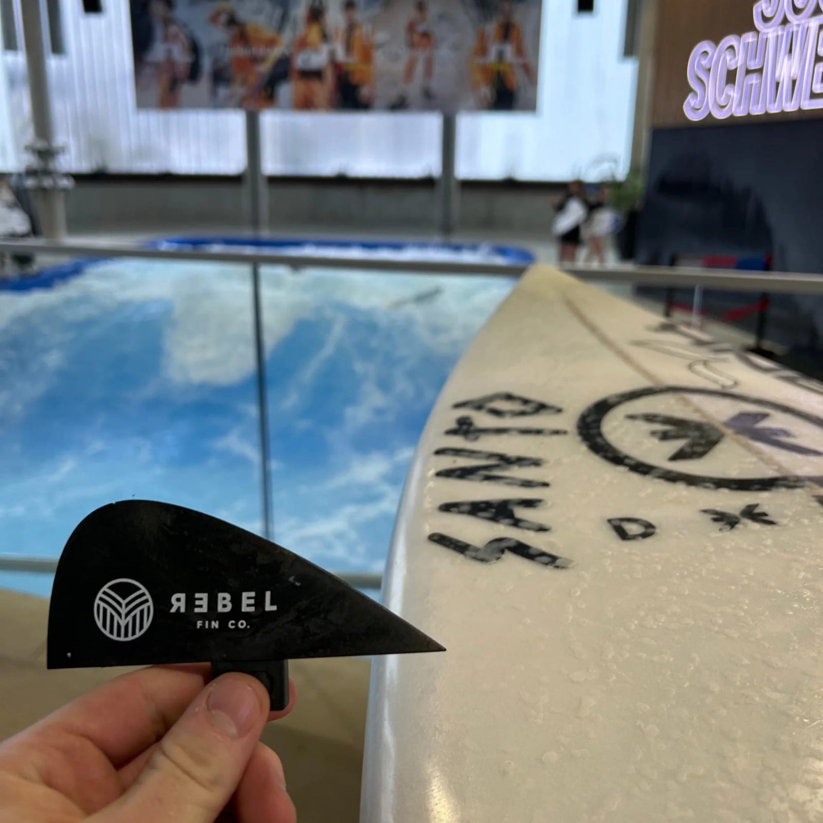 Eine Hand zeigt eine rote Surfari x Rebel Fin - Knubster Finne Glasfaser von Rebel Fin vor einem Santo-Surfbrett, wobei eine Indoor-Welle und ein Surfer-Wandbild die perfekte Kulisse für das Twin-Fin-Setup bilden.