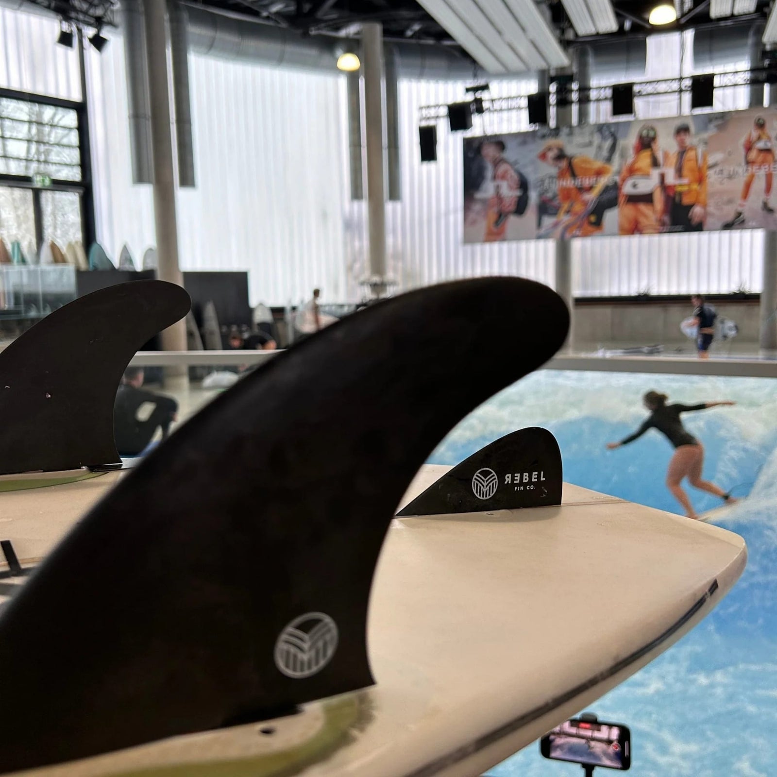 Nahaufnahme der Rebel Fin Surfari x Rebel Fin - Knubster Finne Glasfaser (rot) im Vordergrund, mit einem Surfer, der auf einer Indoor-Welle reitet, in einem modernen Raum mit großen Fenstern, Surfbrettern und einer Wandmalerei zum Thema Sport.