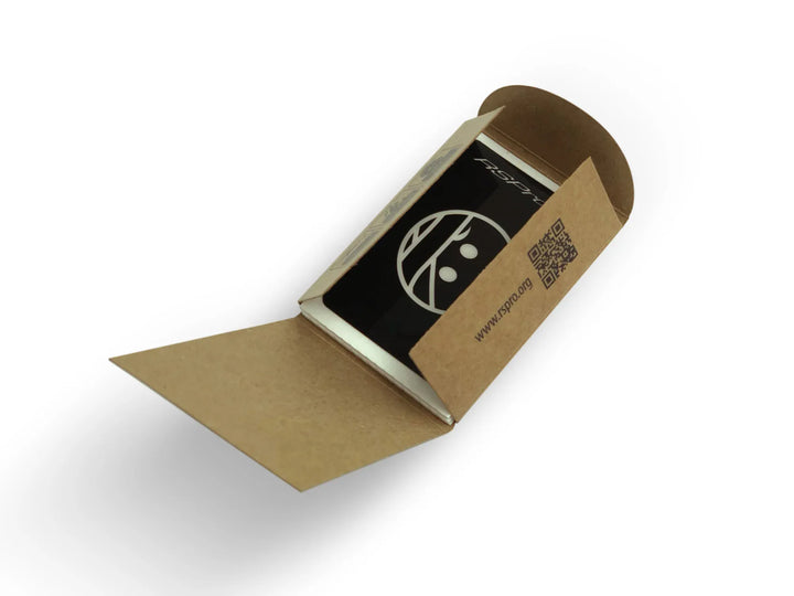 Eine kleine braune Pappschachtel mit einem QR-Code auf der Innenseite des Deckels, teilweise geöffnet, um ein schwarzes RSPro Surfboard Ding Patch (6 Stück) mit einer weißen kreisförmigen Grafik und Text zu enthüllen.