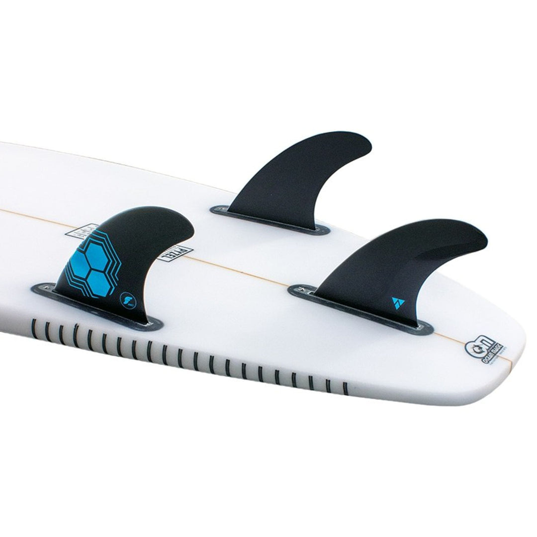 Nahaufnahme eines Channel Islands Surfboard Hecks mit dem futures AM1 Al Merrick Alpha Thruster Finnen Set in schwarz. Das weiße Board hat eine dünne orangefarbene Mittellinie und blau-schwarze Grafiken in der Nähe der Finnenkästen.