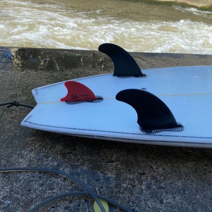 Eine Nahaufnahme eines Surfbretts mit dem Surfboard Finnen - Twin Set M FCSI von Rapid Surf Shop - zwei schwarze Finnen und eine rote Mittelfinne - liegt auf Beton im bewegten Wasser, die Leash ist befestigt, um dieses präzise FCSI Twin Set hervorzuheben.