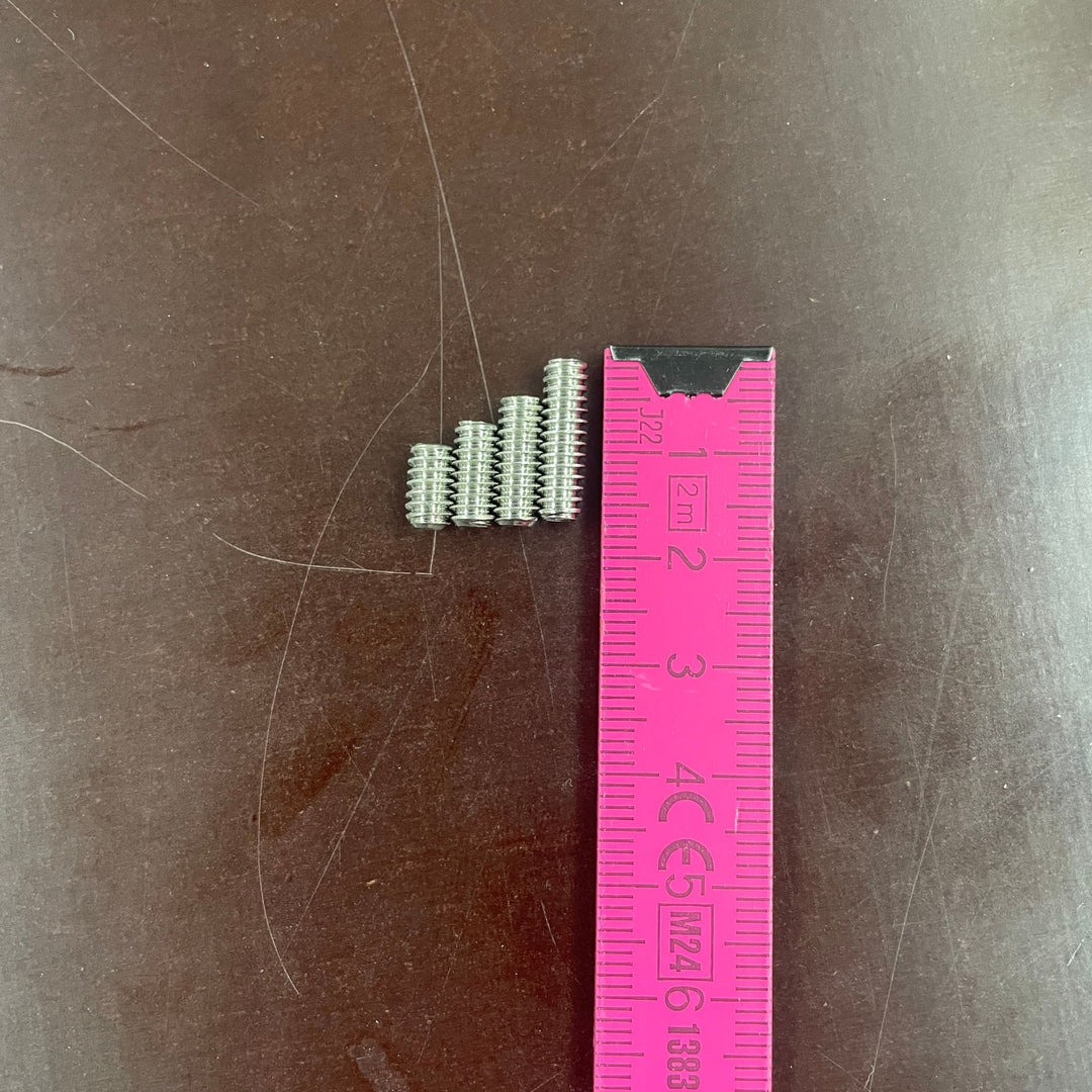 Sechs kleine Finnenschrauben aus Metall aus dem Rapid Surf Shop Finnenschrauben-Set sind neben einem rosa 3 cm Lineal auf einer dunkelbraunen Fläche aufgereiht. Jede ist etwa 1 cm lang, perfekt für FCS Finnenboxen-Setups.