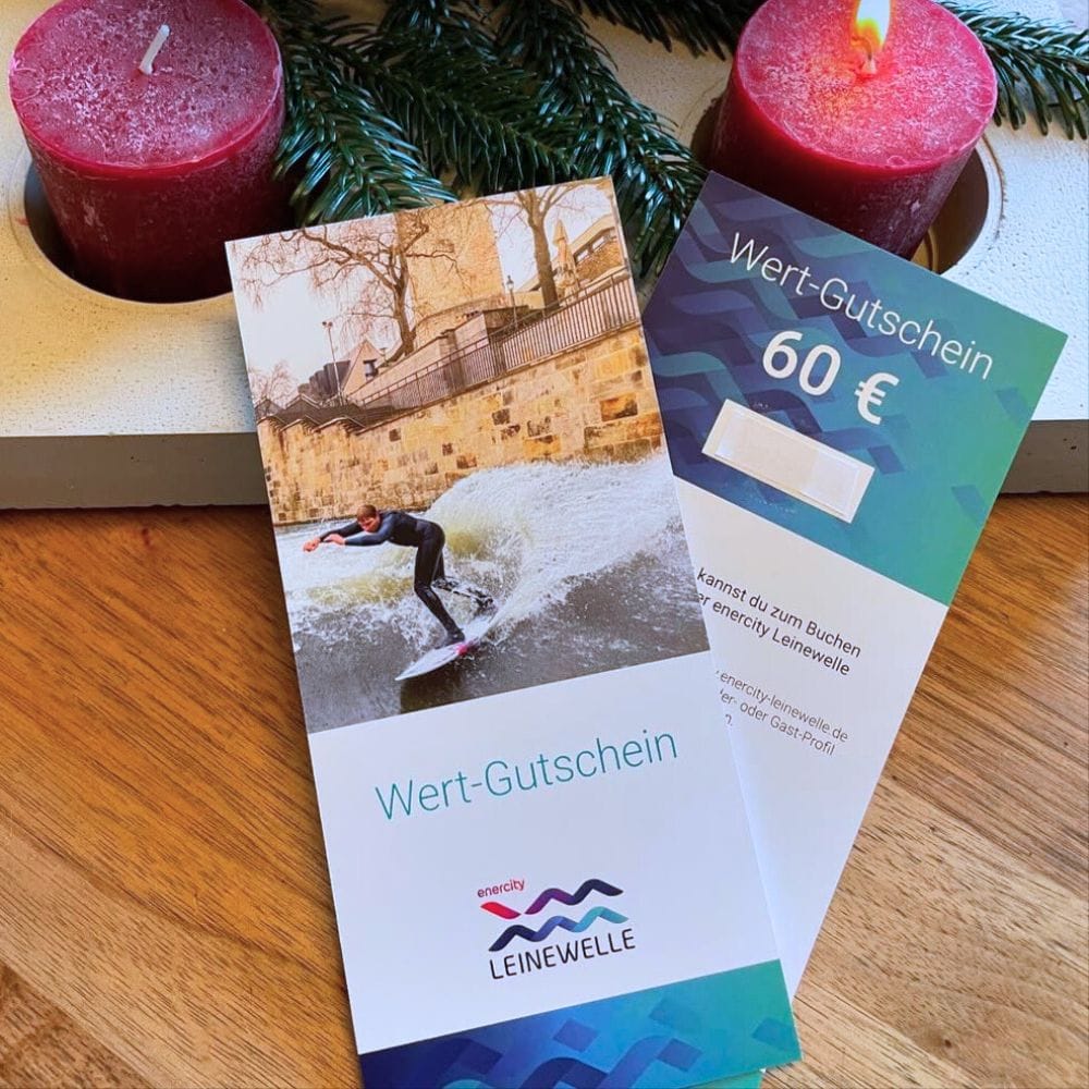 Ein Leinewelle Surf-Gutschein von Rapid Surf Shop im Wert von 60 Euro liegt auf einem Holztisch neben roten Kerzen und Tannenzweigen und zeigt ein surfen Hannover Flusswelle Foto. Ein ideales Print@Home-Geschenk für Surf-Fans.