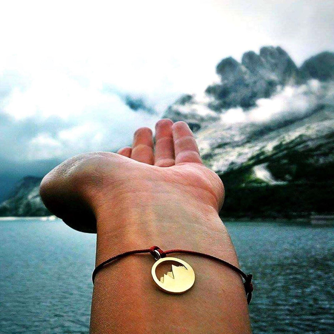 Eine Hand, die das Silver+Surf Silber Schmuck Berg 3D vergoldet Armband mit einem runden, vergoldeten 925er Sterling Silber Anhänger trägt, hält über Wasser mit Bergen und Wolken im Hintergrund.